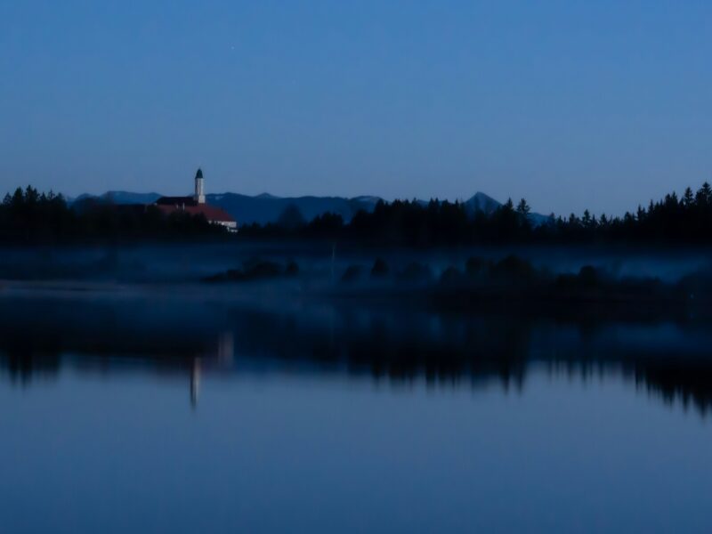 Calm lake reflects dark forest and distant building
